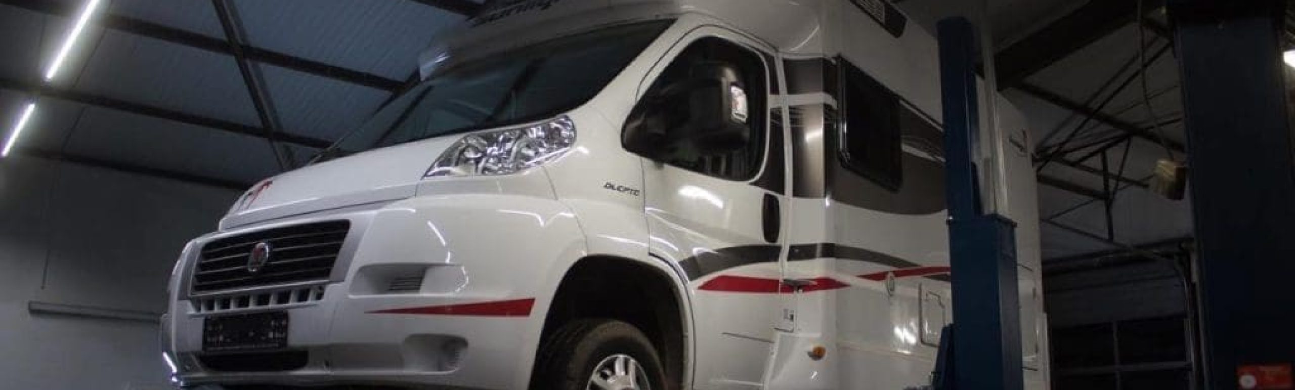 Ein weißes Wohnmobil wird mit einer hydraulischen Hebebühne in einer Garage mit Industriebeleuchtung angehoben. Ein weiterer weißer Lieferwagen ist im Hintergrund mit geöffneter Motorhaube geparkt.