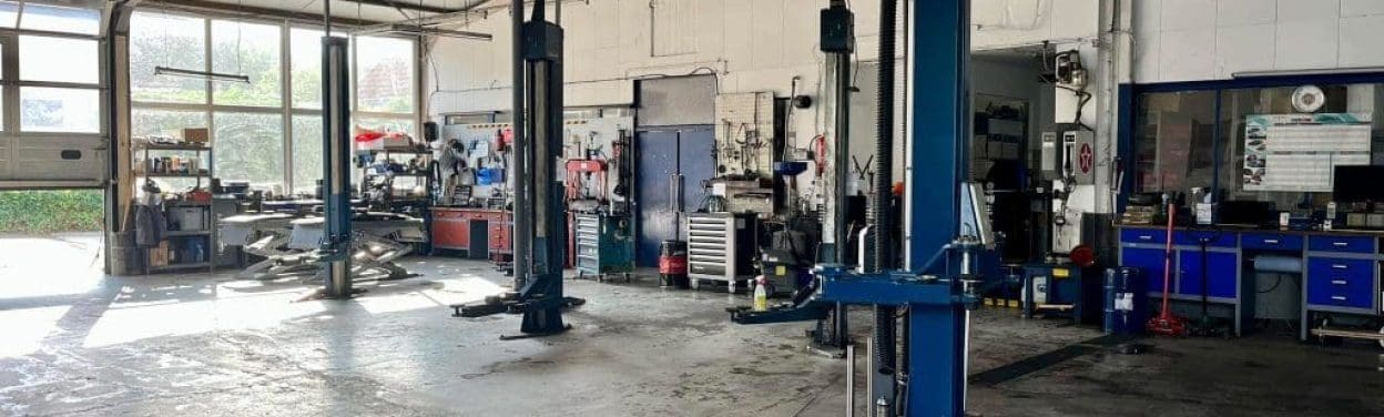 Eine geräumige Autowerkstatt mit Hebebühnen, Werkbänken, verschiedenen Werkzeugen und Geräten. Der Boden ist hell, und durch große Fenster fällt natürliches Licht ein. Die Wände sind weiß, und es gibt ein Schild in deutscher Sprache.