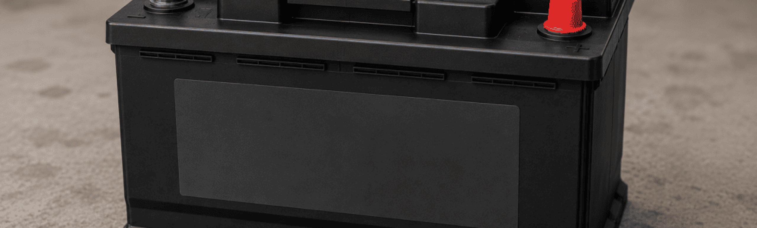 Eine schwarze, defekte Autobatterie mit zwei Polen, von denen einer mit einer roten Kappe abgedeckt ist, liegt auf einem Betonboden. Die Batterie ist rechteckig, weist geprägte Details auf und hat auf der Vorderseite einen leeren Etikettenbereich – das Erkennen von Anzeichen kann hier bei der Hilfe helfen.